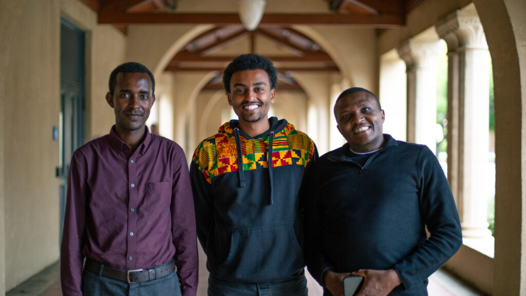 SASH scholars Ombeva and Nahom stand with a Stanford student under Stanford arches on a sunny day